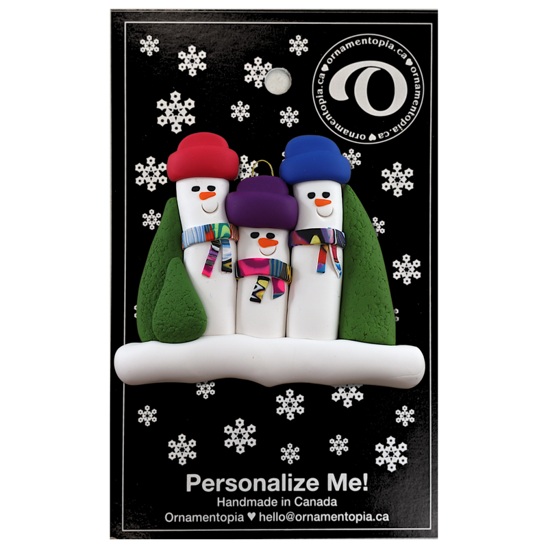 Family ornament with 3 snowmen in colourful scarves and hats, hemmed by Christmas trees, shown on display backing card.