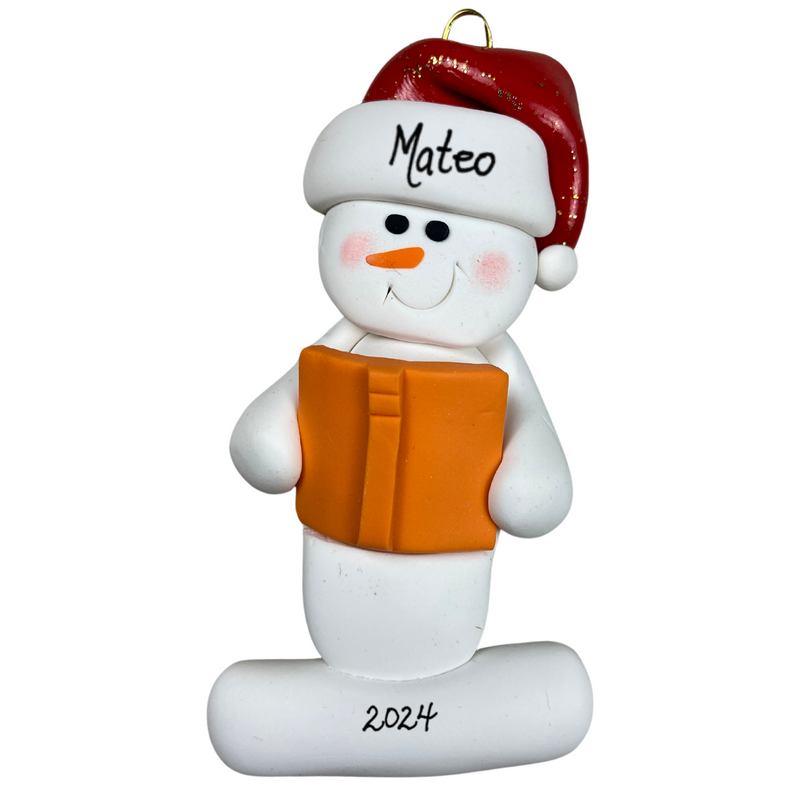Ornament of white snowman in red hat holding an orange book, personalized with name.