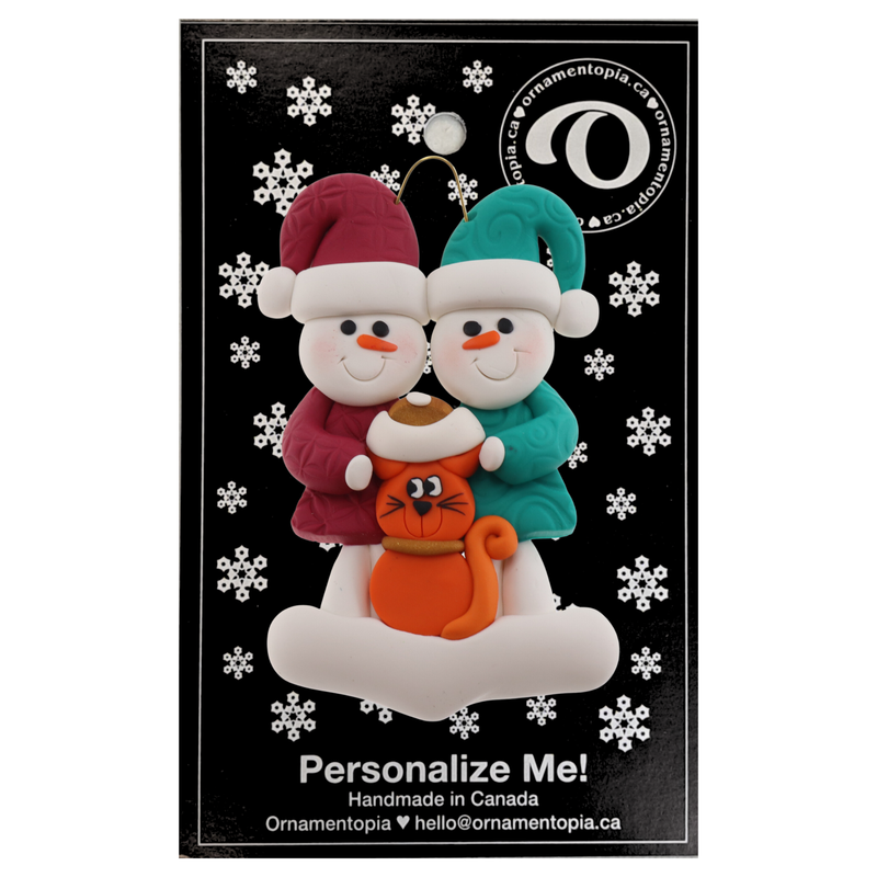 Ornament of snowman couple in red and green shirts and hats with a orange cat in Christmas hat, on a display backing card.