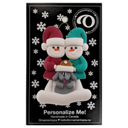Ornament of snowman couple in red and green shirts and hats with grey dog in Christmas hat, on display backing card.