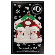 A couple’s ornament of two small white snowmen beside a festive brown house, on display backing card.
