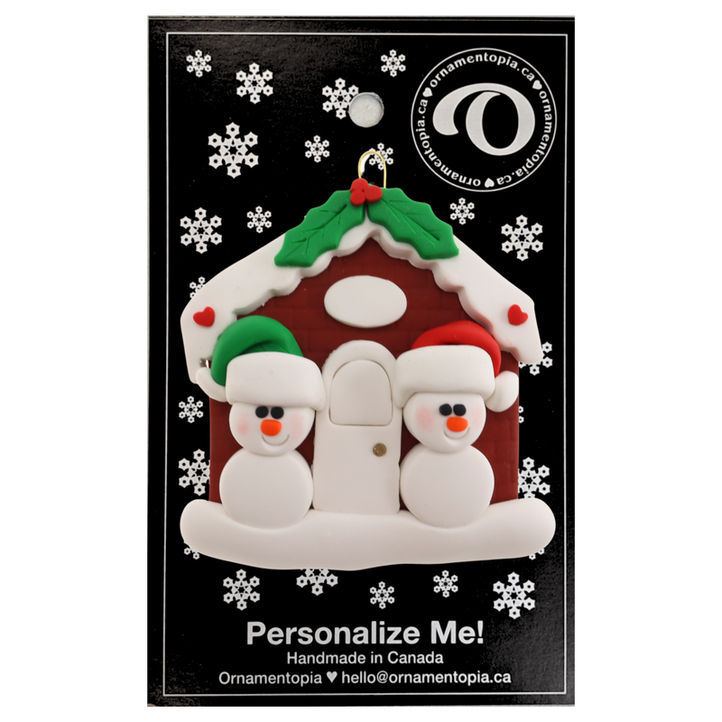 A couple’s ornament of two small white snowmen beside a festive brown house, on display backing card.