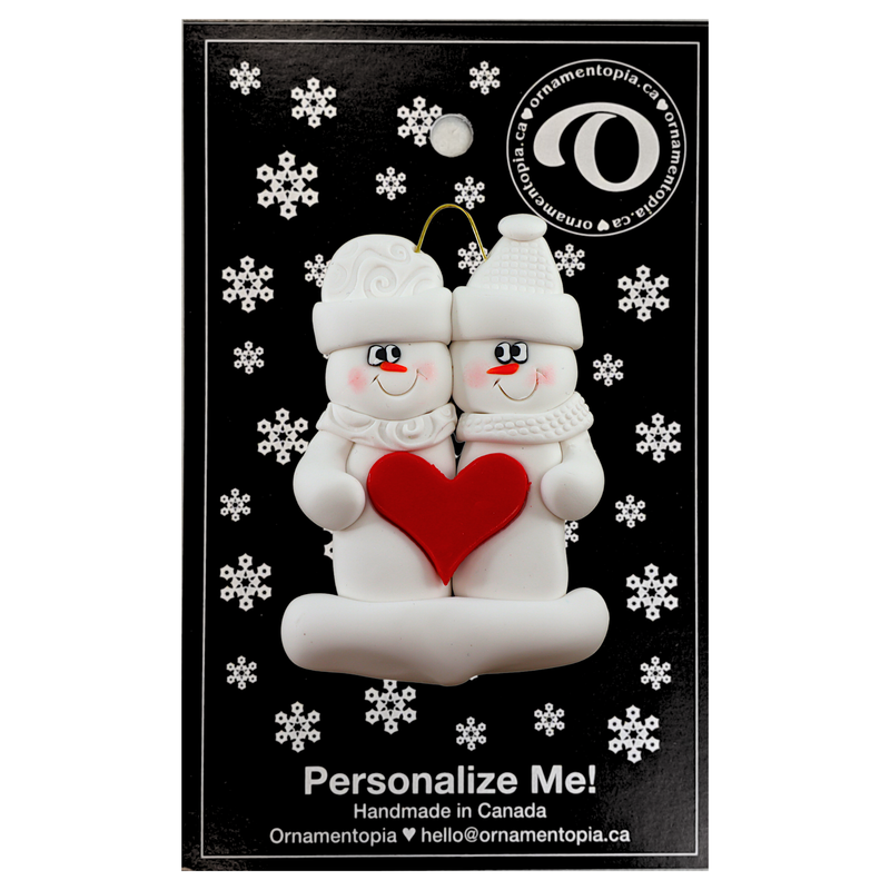A couple’s ornament of two white snowmen holding a red heart together, on display backing card.