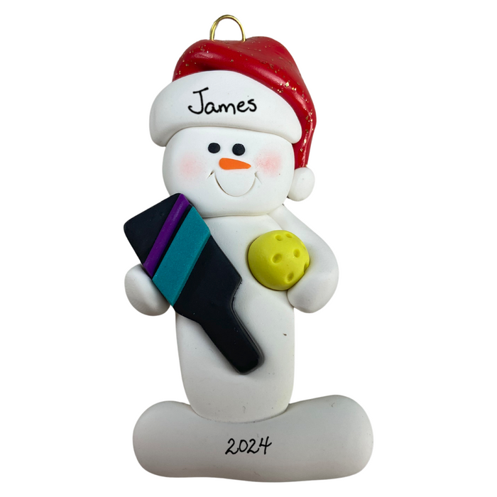 Ornament of white snowman in red hat holding a pickleball paddle and yellow pickleball, personalized with name.