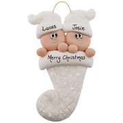 Ornament with twin infant heads in white glitter hats poking out of a white glitter stocking, personalized with name.