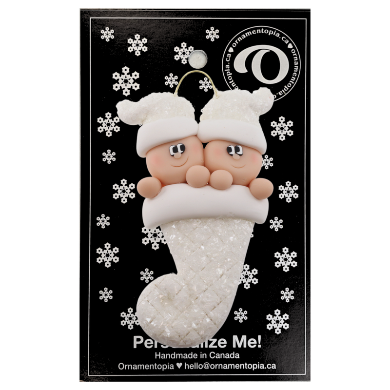 Ornament with twin infant heads in white glitter hats poking out of a white glitter stocking, on display backing card.