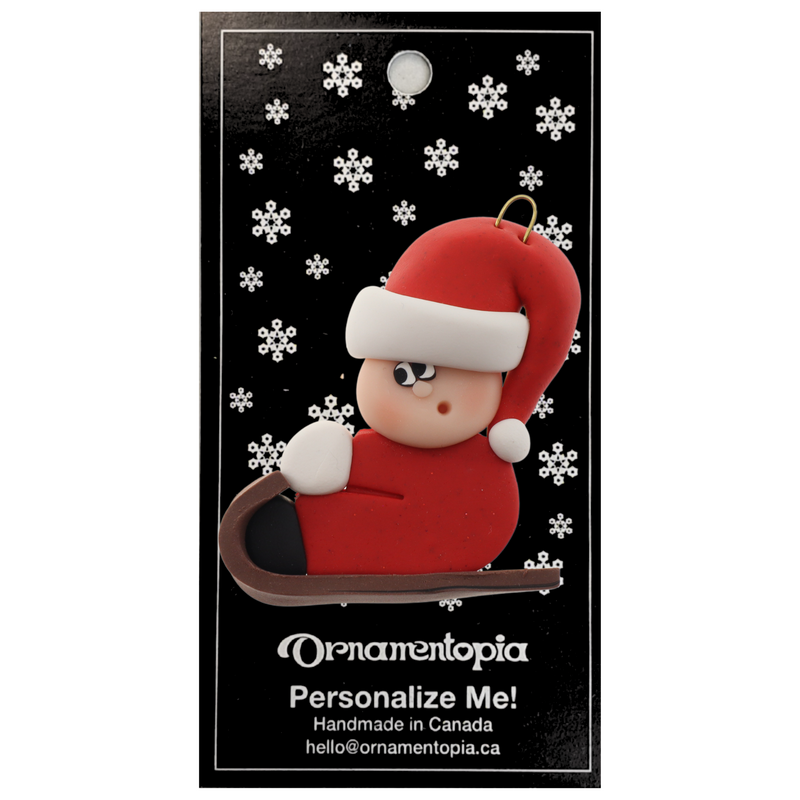 Small figure in red outfit and festive hat riding on brown toboggan ornament, on display backing card.