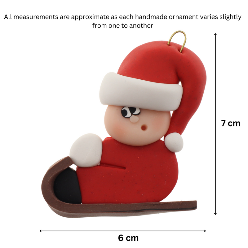 Small figure in red outfit and festive hat riding on brown toboggan ornament, graphed to show size (6x7cm).