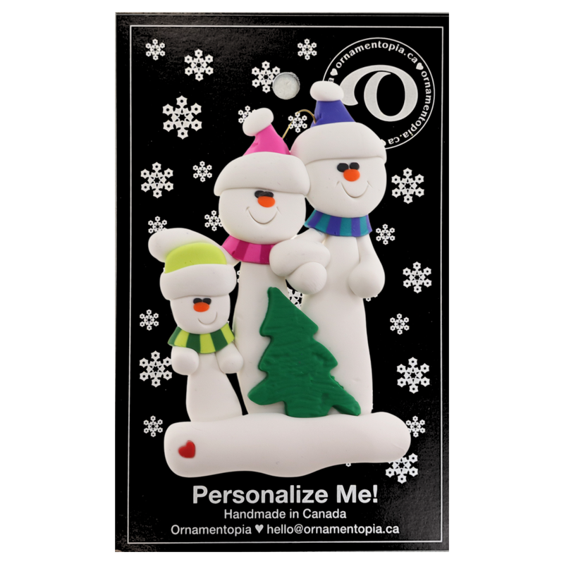 Family ornament with 3 white snowmen in bright scarves, with green Christmas tree, on display card backing.