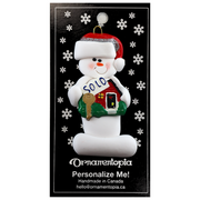 Ornament of white snowman in red hat holding a sign with a house saying “sold”, on display backing card.