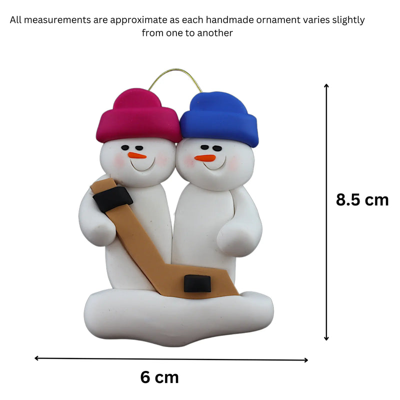 Family ornament with 2 white snowmen in bright hats, holding a hockey stick, graphed to show size (6x8.5cm).