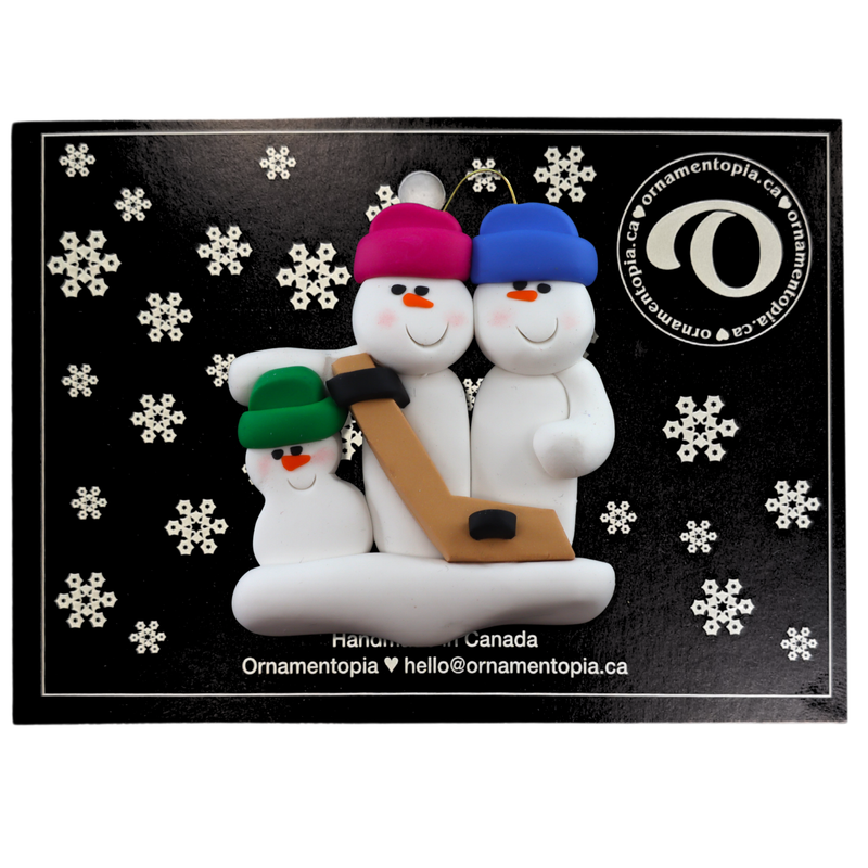 Family ornament with 3 white snowmen in bright hats, holding a hockey stick, on display backing card.