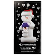 Ornament of white snowman in red hat holding smaller snowman, on display backing card.