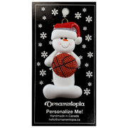 Ornament of white snowman in red hat holding a basketball, on display backing card.