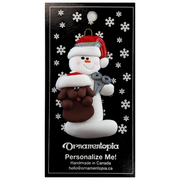 Ornament of white snowman in red hat holding a brown dog and a pair of scissors, on display backing card.