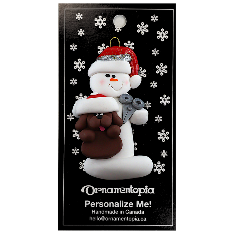 Ornament of white snowman in red hat holding a brown dog and a pair of scissors, on display backing card.