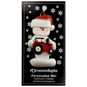 Ornament of white snowman in red hat holding a red tractor, on display backing card.