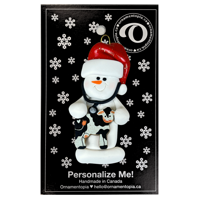 Ornament of white snowman in red hat holding a cow and stethoscope, on display backing card.