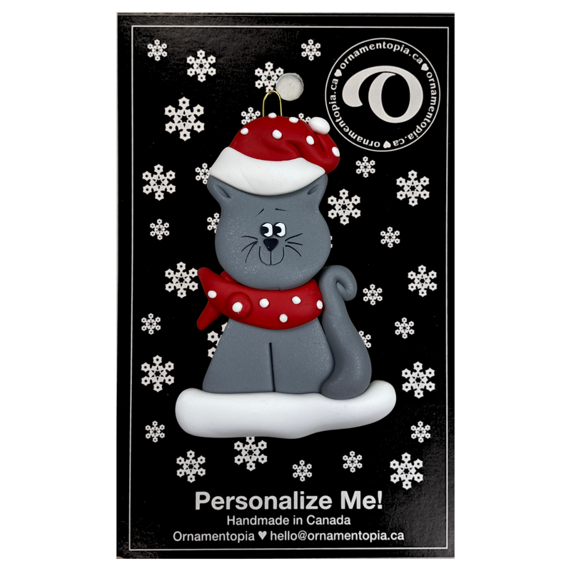 Ornament of grey cat in red and white polka dot Christmas hat and scarf, on display backing card.