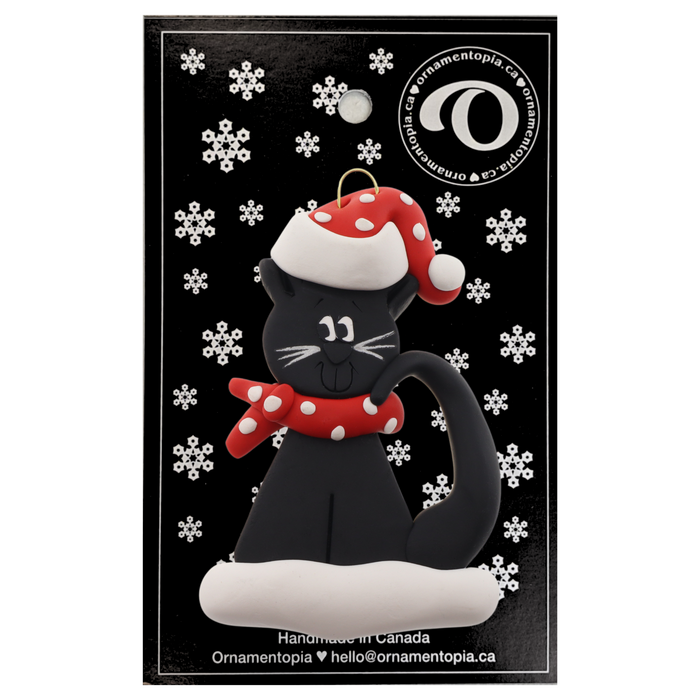 Ornament of black cat in red and white polka dot Christmas hat and scarf, on display backing card.