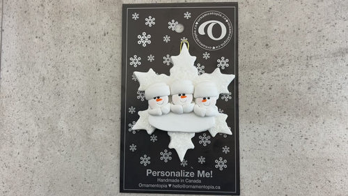 Family ornament with 3 white snowmen stacked on top of a glitter white snowflake, shown on backing card and Christmas tree.