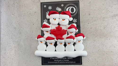Family ornament with 9 white snowmen in red hats, gathered around a red maple leaf, shown on backing card and Christmas tree.