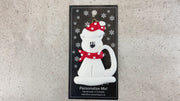 Ornament of white cat in red and white polka dot Christmas hat and scarf, shown on backing card and Christmas tree.