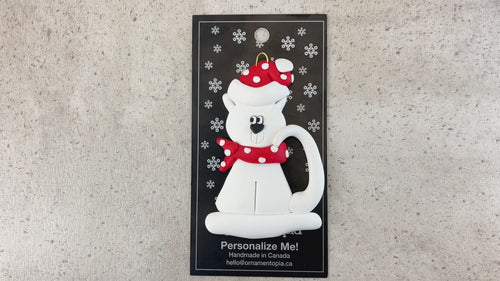Ornament of white cat in red and white polka dot Christmas hat and scarf, shown on backing card and Christmas tree.