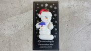 Ornament of white snowman in red hat holding smaller snowman, shown on backing card and Christmas tree.