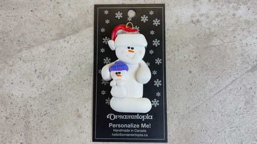 Ornament of white snowman in red hat holding smaller snowman, shown on backing card and Christmas tree.