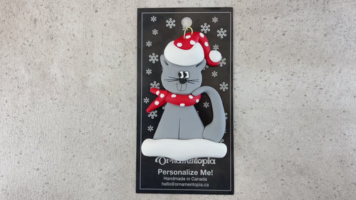 Ornament of grey cat in red and white polka dot Christmas hat and scarf, shown on backing card and Christmas tree.
