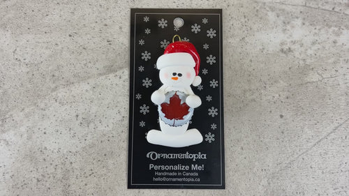 Ornament of white snowman in red hat holding a beer bottle cap, shown on backing card and Christmas tree.
