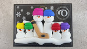 Family ornament with 6 white snowmen in bright hats, holding a hockey stick, shown on backing card and Christmas tree.