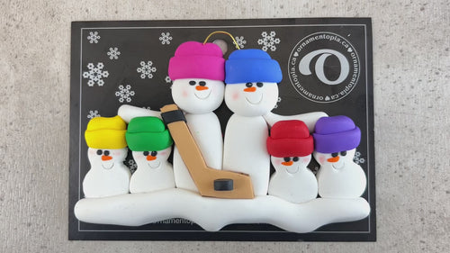 Family ornament with 6 white snowmen in bright hats, holding a hockey stick, shown on backing card and Christmas tree.