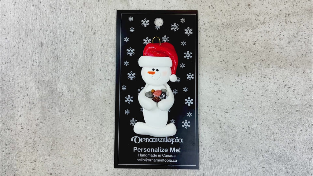 Ornament of white snowman in red hat holding several small rocks, shown on backing card and Christmas tree.