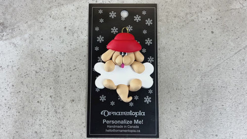 Ornament of tan dog in red hat hanging on a white bone, shown on backing card and Christmas tree.