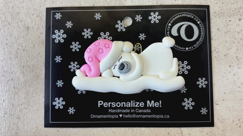 Ornament of white sleeping polar bear baby in a pink hat, shown on backing card and Christmas tree.