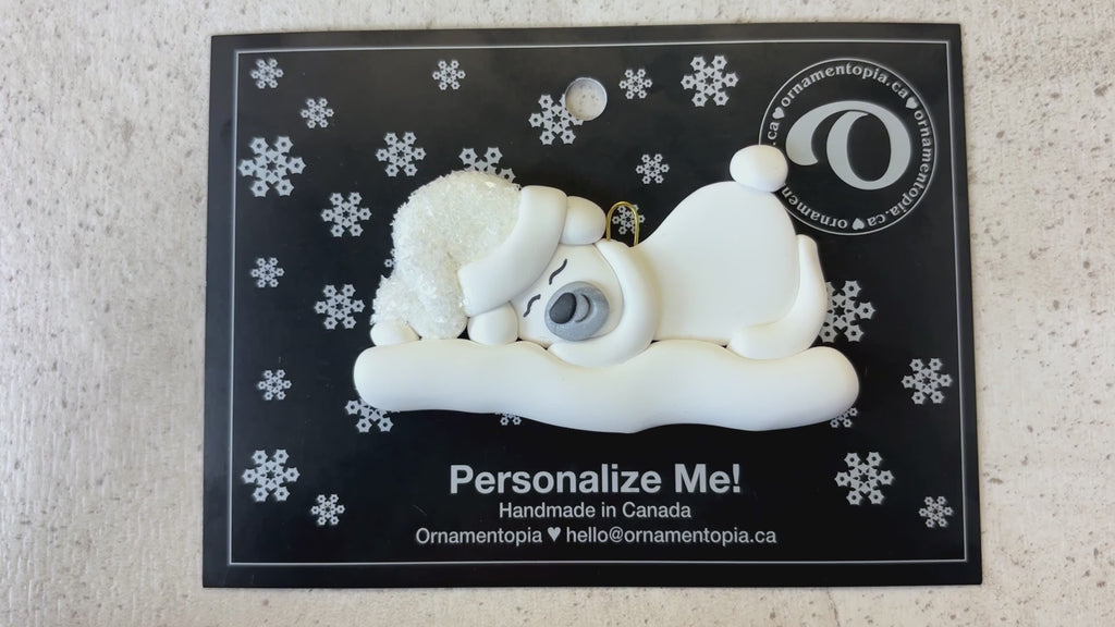 Ornament of white sleeping polar bear baby in a white sparkling hat, shown on backing card and Christmas tree.