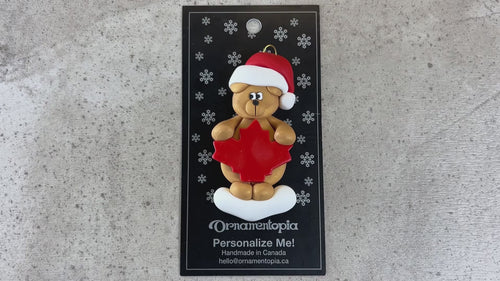 Tan bear in red festive hat with maple leaf ornament, shown on backing card and Christmas tree.