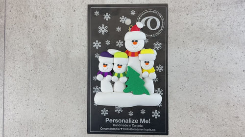 Family ornament with 1 large and 3 small white snowmen in bright scarves, with green Christmas tree, shown on backing card and Christmas tree.