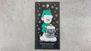 Ornament of snowman in green shirt and hat with grey dog in Christmas hat, shown on backing card and Christmas tree.