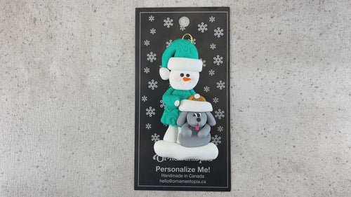 Ornament of snowman in green shirt and hat with grey dog in Christmas hat, shown on backing card and Christmas tree.