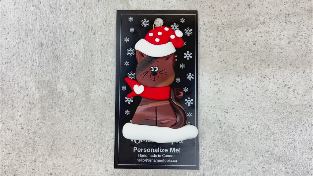 Ornament of brown cat in red and white polka dot Christmas hat and scarf, shown on backing card and Christmas tree.