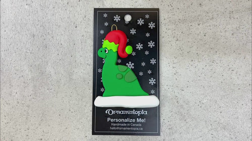 Green dinosaur wearing red festive hat ornament, shown on backing card and Christmas tree.