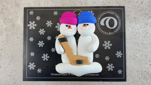 Family ornament with 2 white snowmen in bright hats, holding a hockey stick, shown on backing card and Christmas tree.