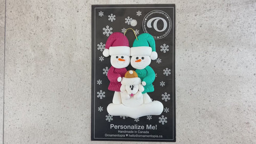 Ornament of snowman couple in red and green shirts and hats with white dog in Christmas hat, shown on backing card and Christmas tree.