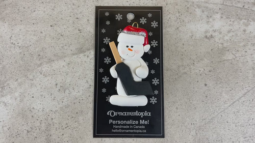 Ornament of white snowman in red hat holding a goalie stick, shown on backing card and Christmas tree.