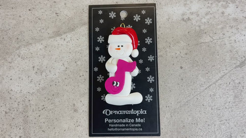 Ornament of white snowman in red hat holding a purple musical note, shown on backing card and Christmas tree.