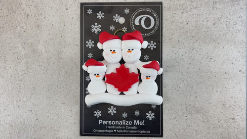 Family ornament with 4 white snowmen in red hats, gathered around a red maple leaf, shown on backing card and Christmas tree.