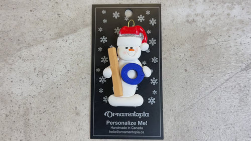Ornament of white snowman in red hat holding a ringette stick and blue ring, shown on backing card and Christmas tree.